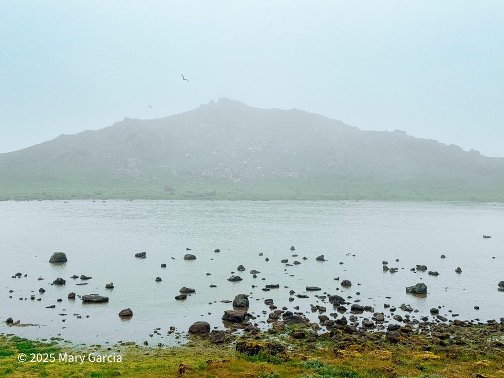 Kittiwake Lake with scattered rocks in the water and a large hill in the background, St. Paul Island, Alaska.