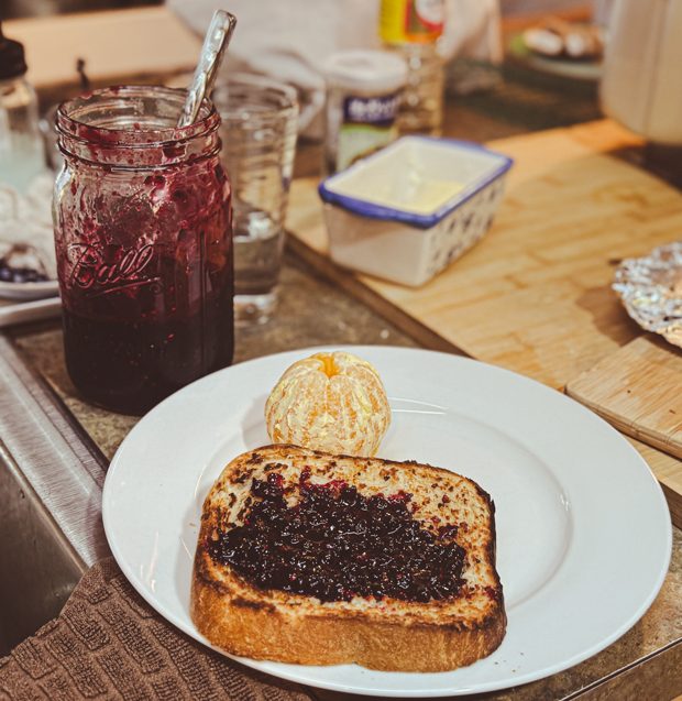 Plate with toast spread with dark purple mossberry jam, served with a peeled clementine, next to a jar of homemade jam.