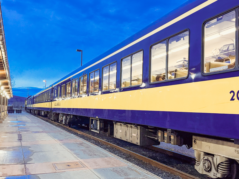 Blue and yellow Alaska Railroad train at a station platform under a twilight sky.
